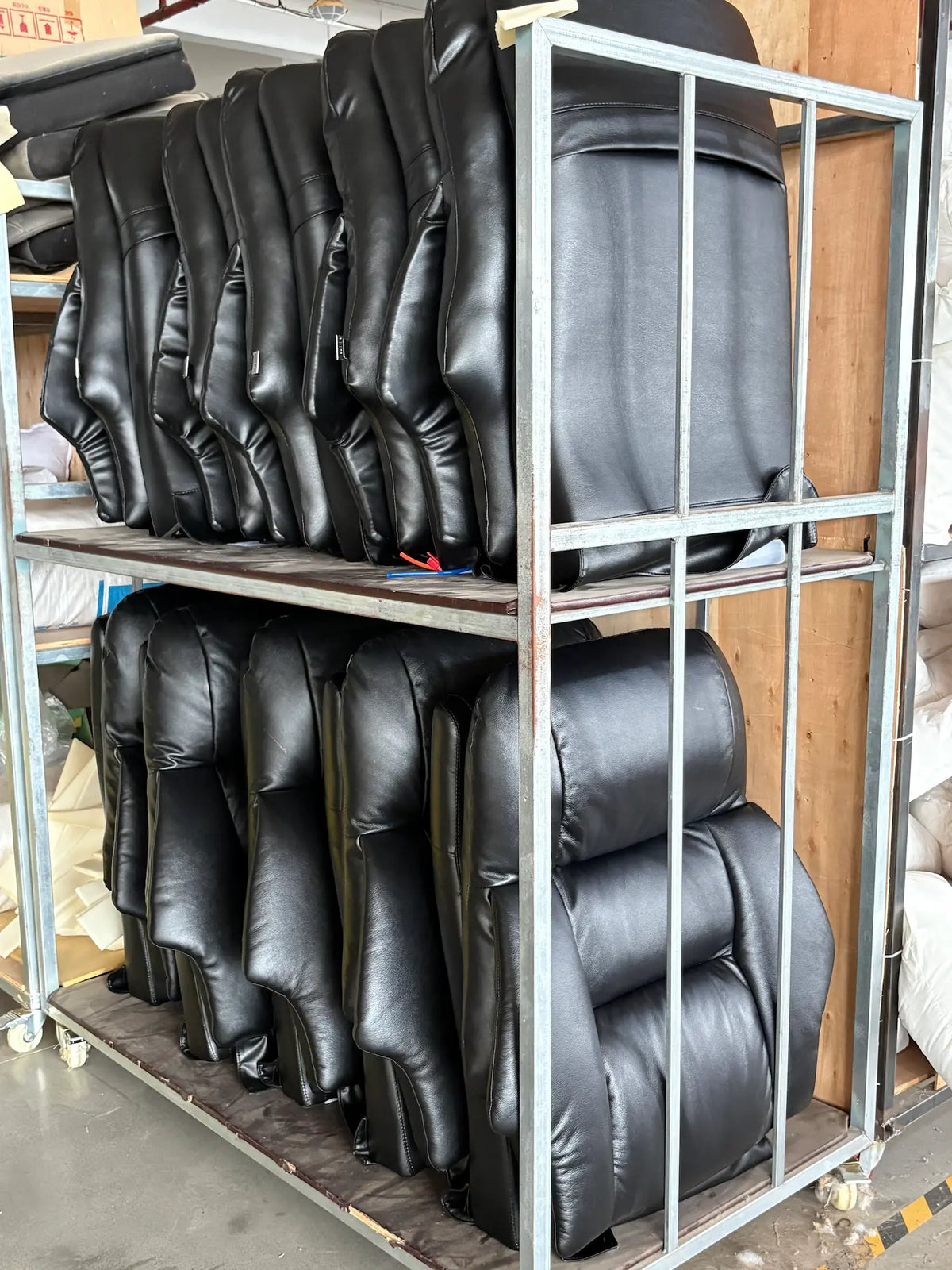 Several black leather or faux-leather chair backrests or upholstered seat cushions stacked on a metal shelving unit in a warehouse or workshop setting.