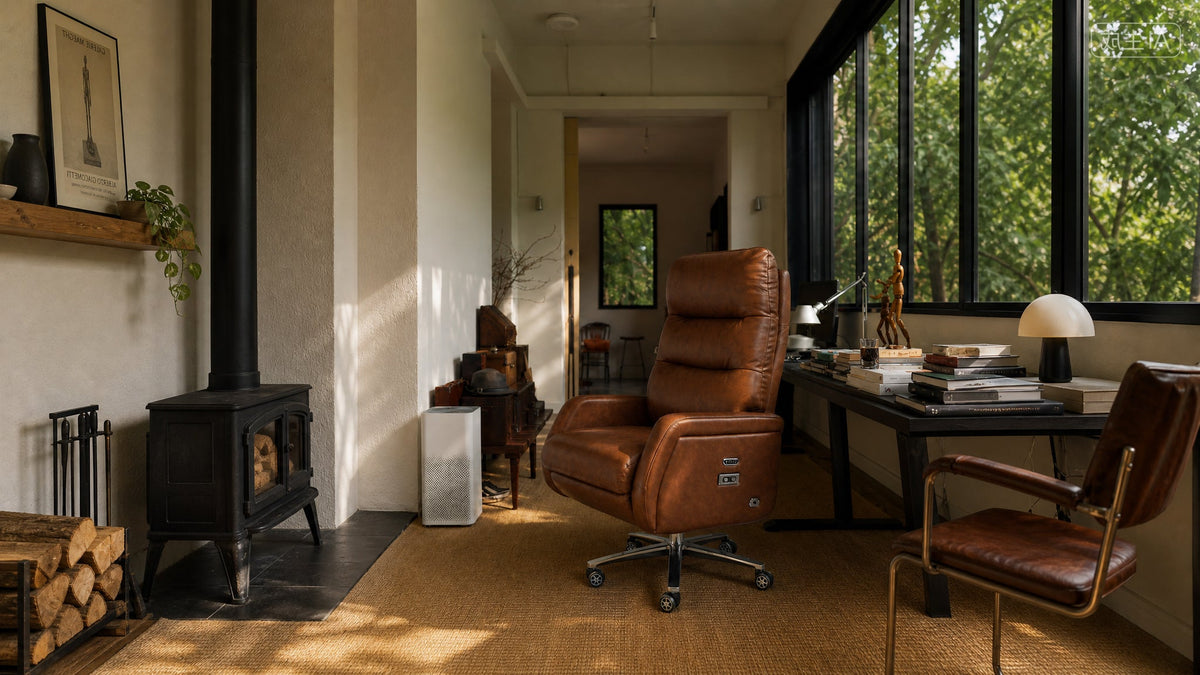 A warm brown leather office chair with a five-star swivel base is positioned in a sunlit home office interior next to a desk and large windows.