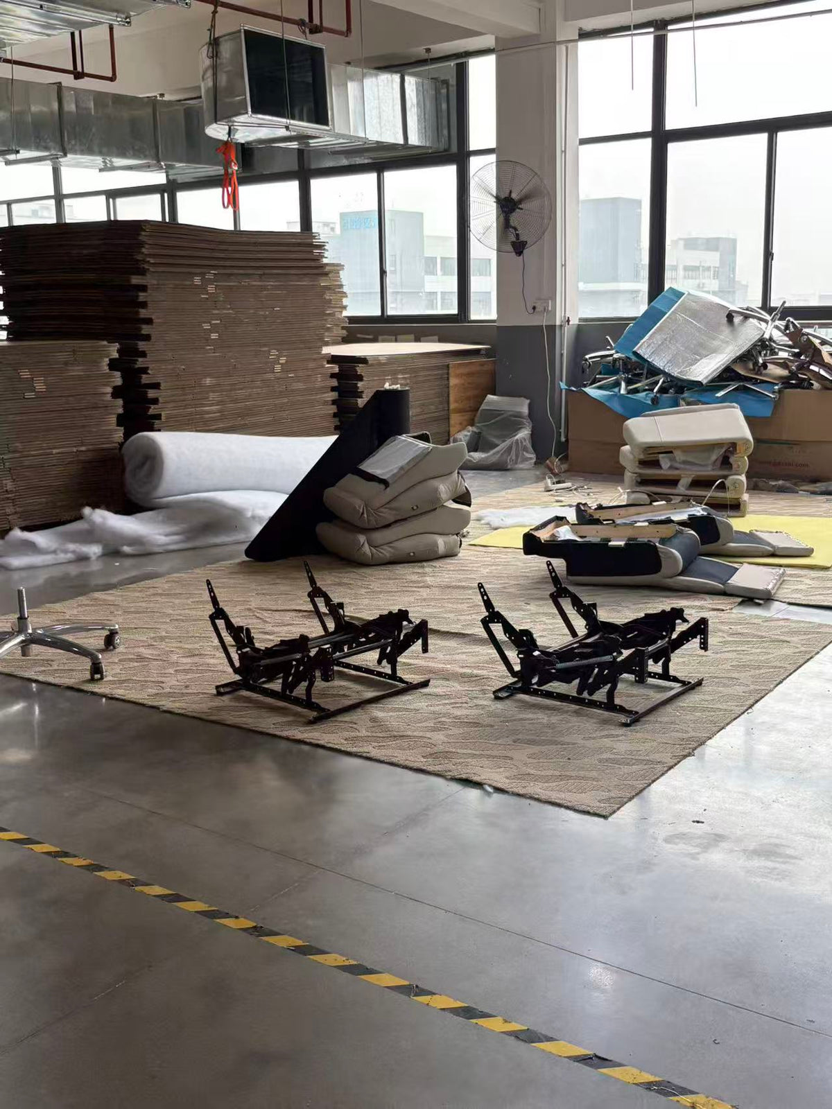 A warehouse or assembly area floor showing two black metal chair frames on a beige rug, surrounded by stacks of cardboard, upholstered cushions, and other furniture parts. Large industrial windows and ceiling ducts are visible in the background.