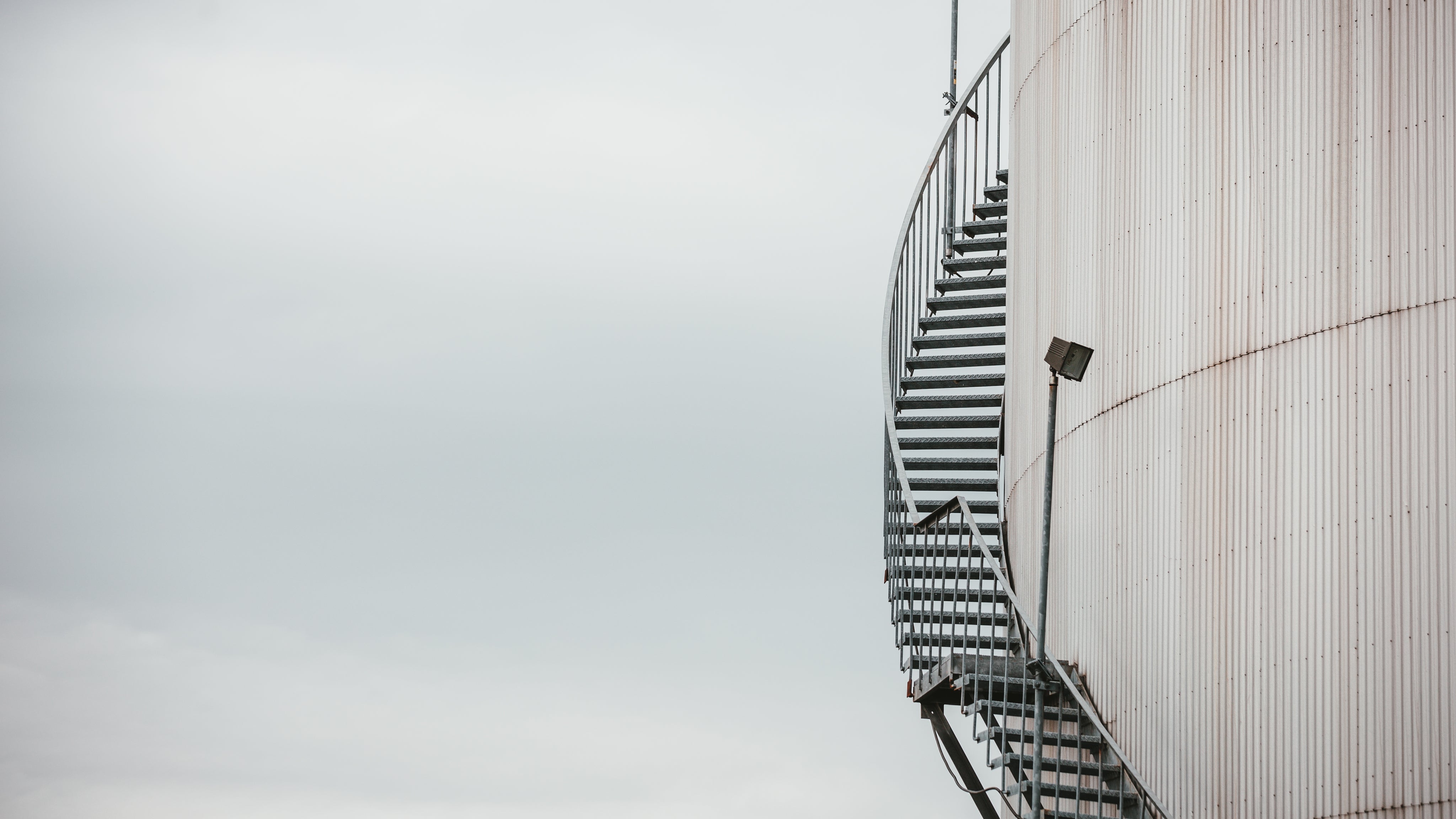 A curved metal spiral staircase with a black railing attached to a large white cylindrical tank under an overcast gray sky.