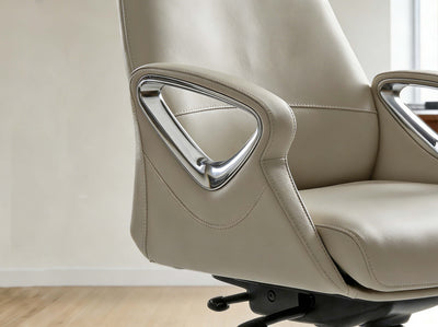 Close-up of a beige leather office chair with a sculpted chrome side armrest, set against a minimalist light beige wall and light wood floor.