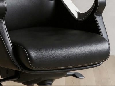 Close-up of a black faux leather office chair seat cushion with visible stitching, showing part of the armrest and a light wood floor in the background.