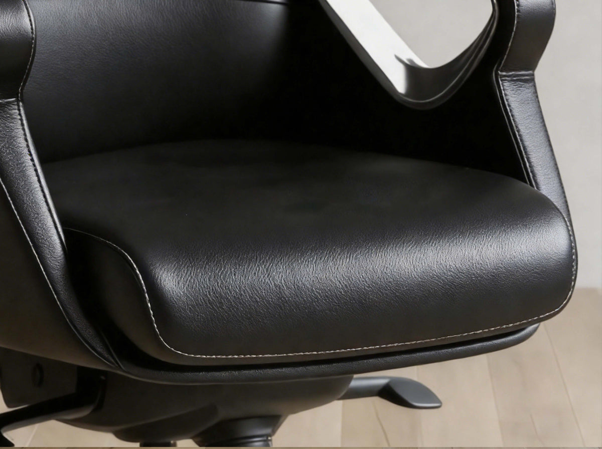 Close-up of a black faux leather office chair seat cushion with visible stitching, showing part of the armrest and a light wood floor in the background.