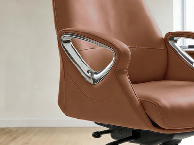 Close-up of a modern office chair upholstered in warm brown leather with polished chrome triangular armrest accents, shown in a clean indoor setting with light wood flooring and a neutral wall.
